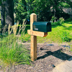 Newly built wooden mailbox stand with black steel mailbox and white address numbers on side. Landscape grass beside mailbox with leafy green trees in background with house glimpsed through tree trunks. Used beside contact form for mailbox installation and repair company, Father and Son Building.