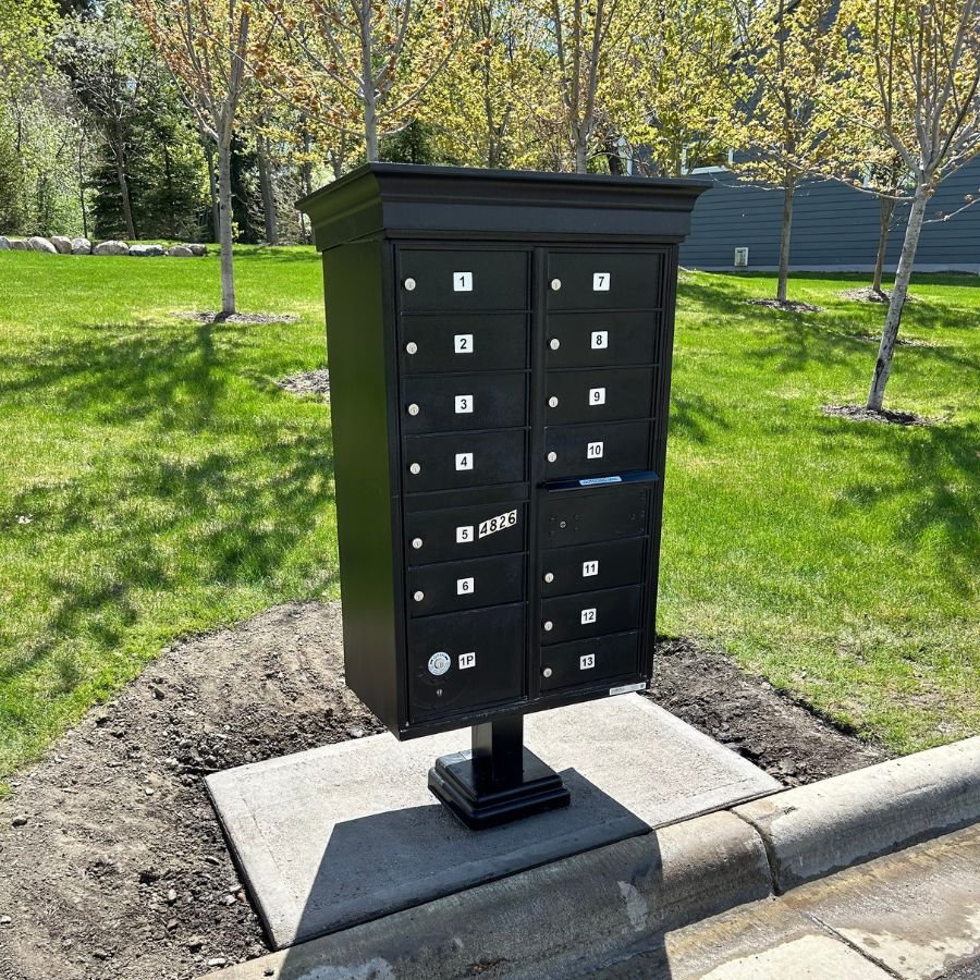 Commercial Mailbox Installation - Black Multi-mailbox on grey concrete foundation with trees and green lawn in background.